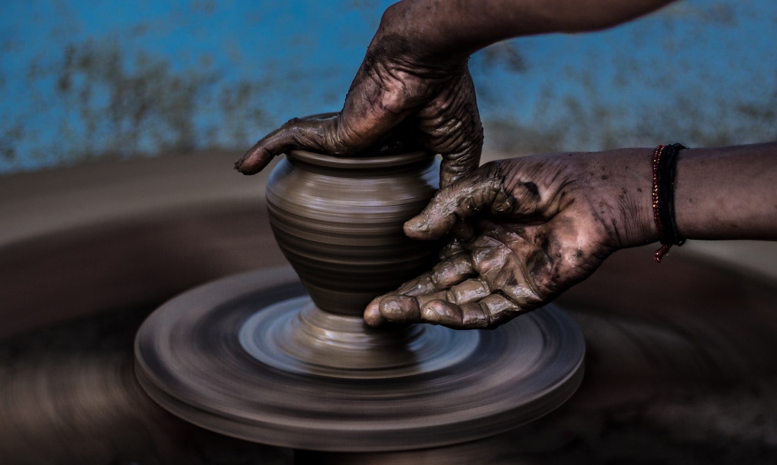 A potter's clay-covered hands shaping a small pot on a spinning wheel
